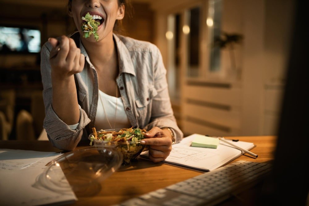 female freelancer using computer eating salad night home min