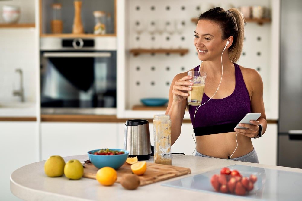 happy athletic woman drinking smoothie while using mobile phone listening music ear phone kitchen min